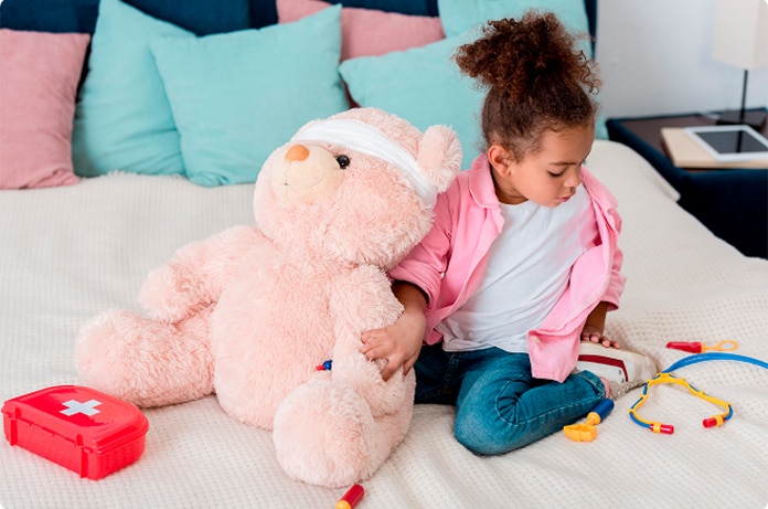 Menor sentado en una cama cuidando a un oso de peluche vendado junto a un estuche médico de juguete