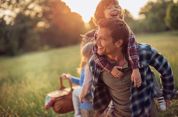 Grupo familiar caminando por un claro de bosque, el adulto lleva a su espalda a un menor sonriente