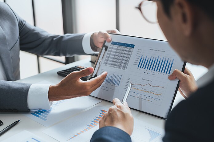 Dos personas revisando gráficos financieros en una tableta mientras señalan distintos datos sobre una mesa con documentos impresos