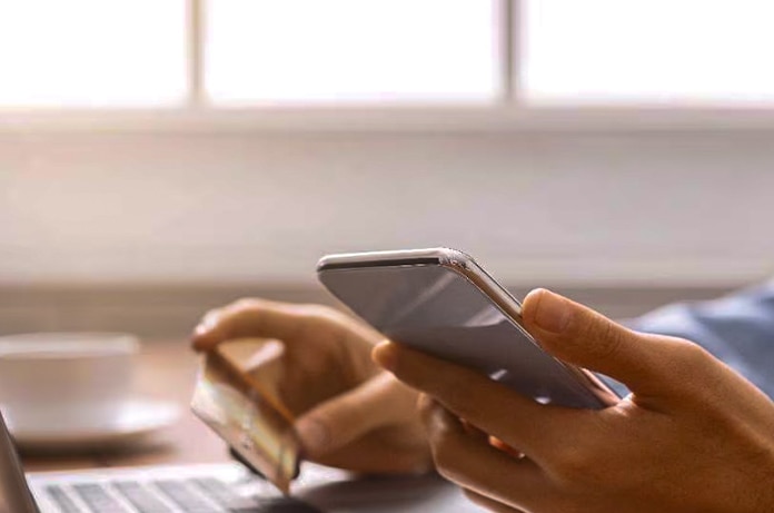 Manos sosteniendo un teléfono móvil y una tarjeta bancaria frente a un ordenador portátil durante una operación de compra online