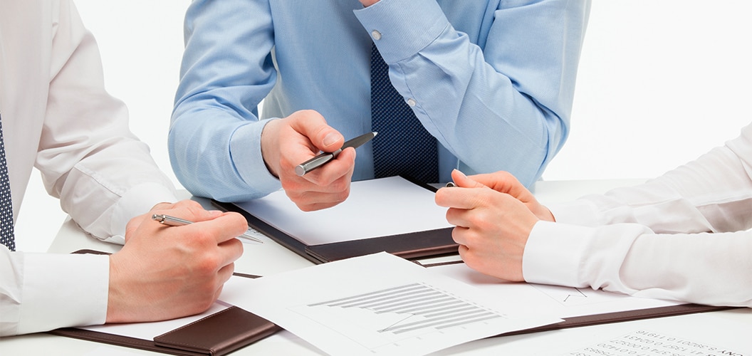 Varias personas reunidas en una mesa revisando documentos impresos y gráficos mientras señalan diferentes puntos con bolígrafos