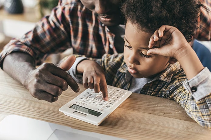Manos de una persona adulta guiando a una criatura mientras pulsa las teclas de una calculadora sobre una mesa de madera